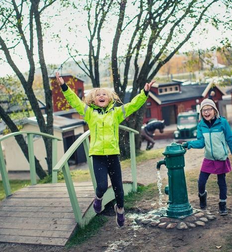 Två barn. Det ena barnet hoppar och det andra barnet har handen på en gammalmodig vattenpump.