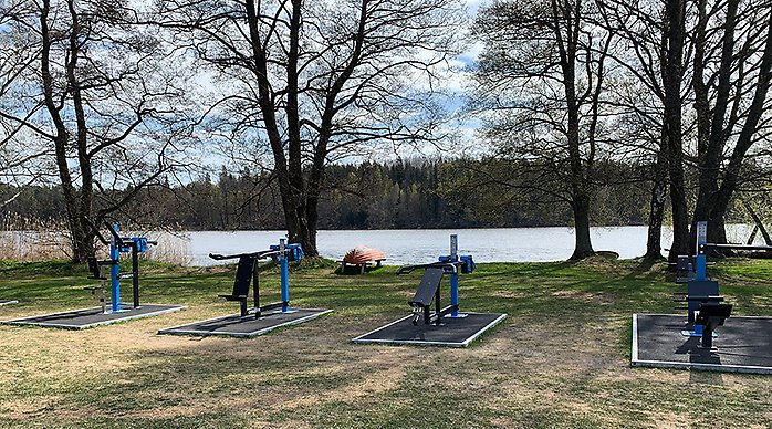 Olika träningsredskap i ett utegym med träd och vatten i bakgrunden.