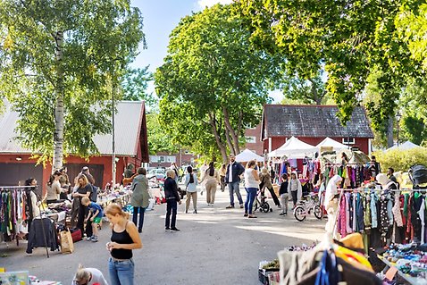 En solig dag vid återbruksloppisen på festivalområdet. Längs gångvägen står bord och klädställningar uppställda med begagnade kläder och prylar. Många människor går runt, pratar och tittar på saker. I bakgrunden syns röda trähus och tält, omgivna av grönskande träd.